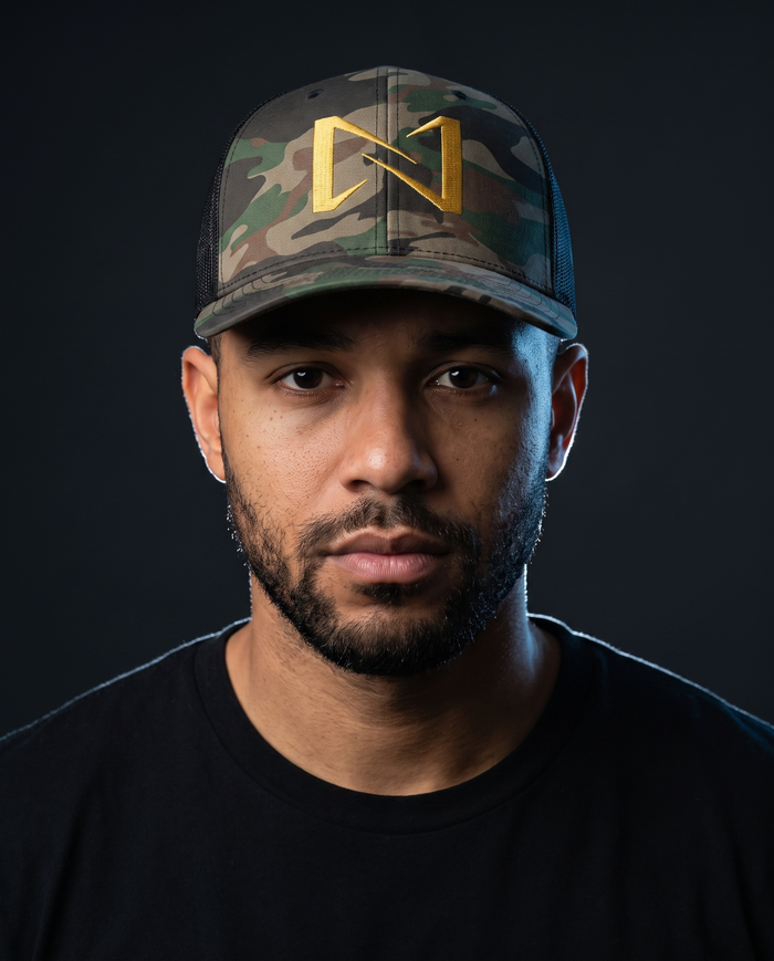 Model wearing the NPRWLR N Logo Camouflage Trucker Hat, front view showing the gold embroidered N on the camo front panel