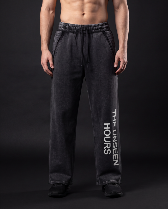 Model wearing the NPRWLR Snow Wash Raw Edge Sweatpants in black, front view showing THE UNSEEN HOURS printed vertically down the leg